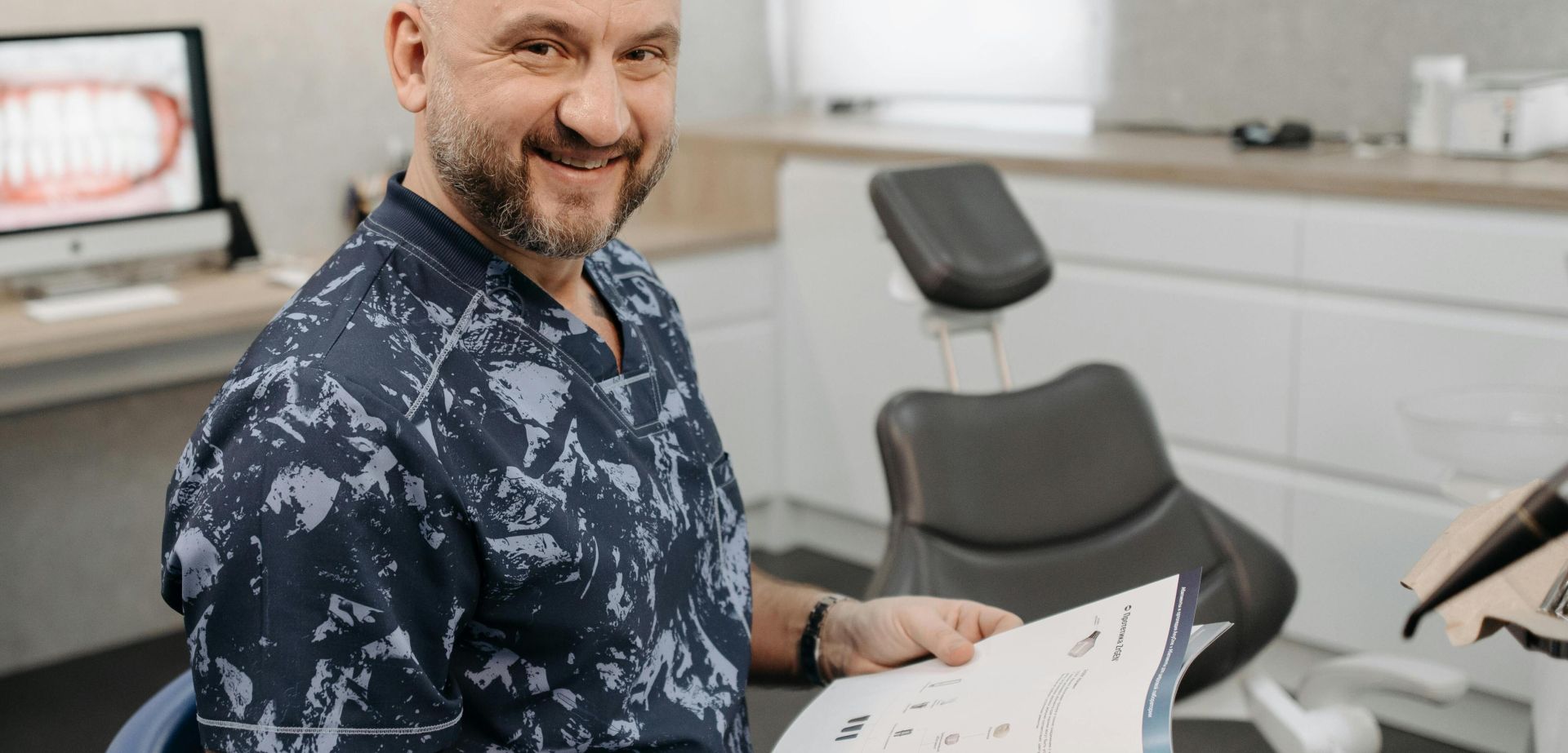 Smiling dentist holding a book, sitting in a dental office with equipment around.