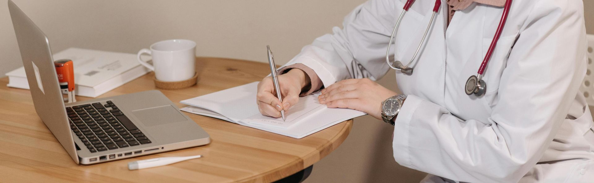 Woman doctor with stethoscope writing at desk with laptop and notes.